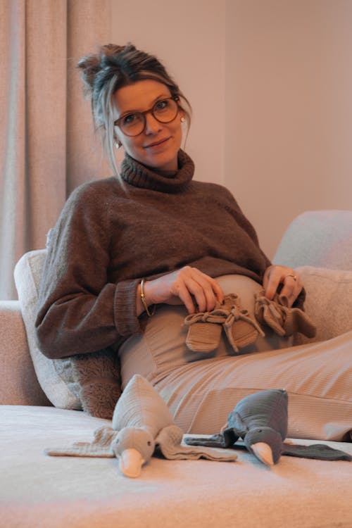 Smiling Pregnant Woman Sitting on the Sofa Holding Baby Shoes