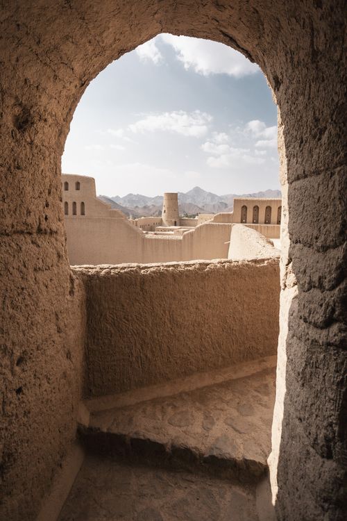 Free View of Nizwa Fort in Oman through a rustic stone archway with desert mountains in the background. Stock Photo