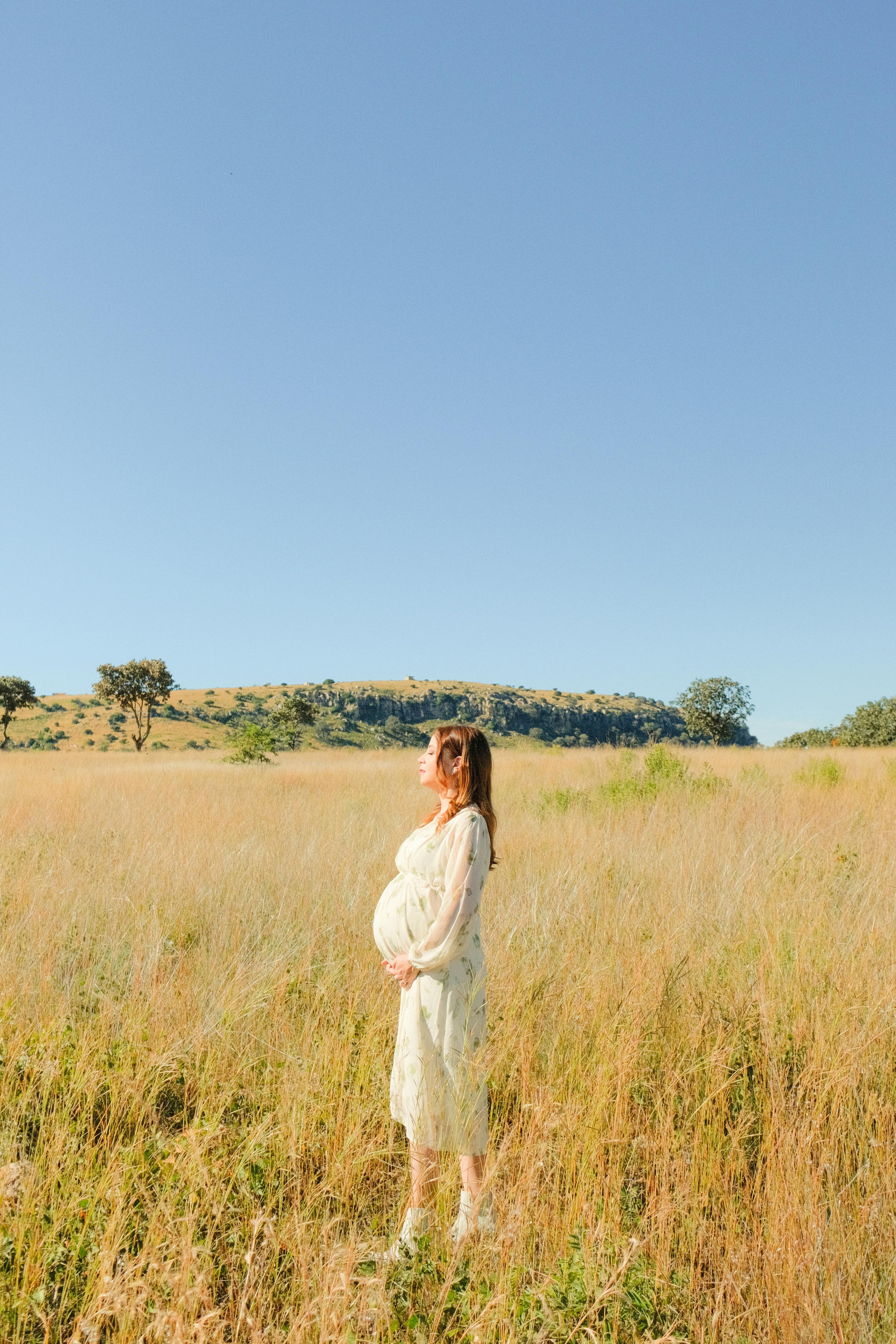 A serene image of a pregnant woman in a white dress standing in a sunlit hayfield during summer.