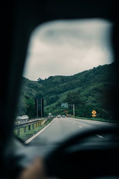 View through car window on a road trip through a lush green landscape, featuring distant hills and cloudy skies.
