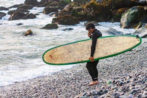 Surfer with a Board Under His Arm Standing on the Pebbles of the Beach