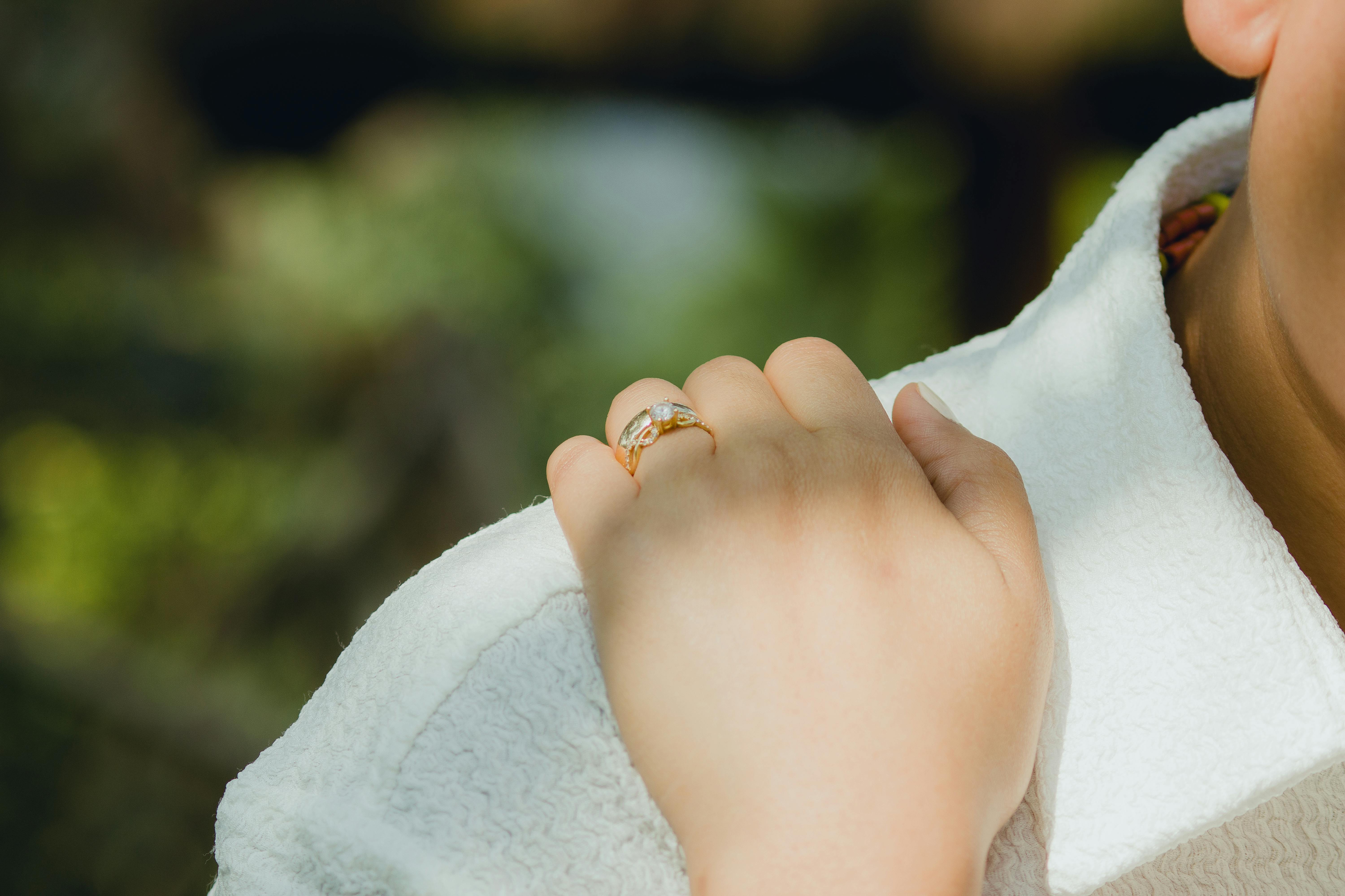 A close-up photograph of a hand with an engagement ring resting on a shoulder outdoors.
