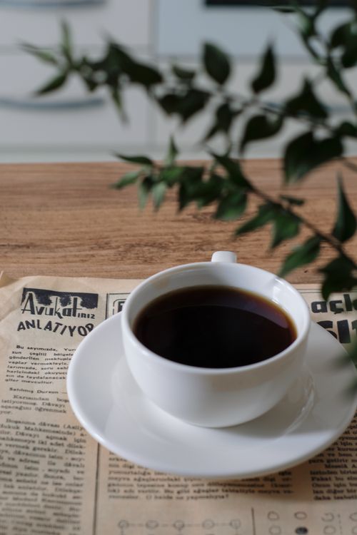 Free A white coffee cup on a saucer with black coffee, placed on a newspaper with green leaves nearby. Stock Photo