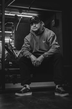 Stylish man with hooded jacket and cap posing in gym, exuding street fashion vibe.