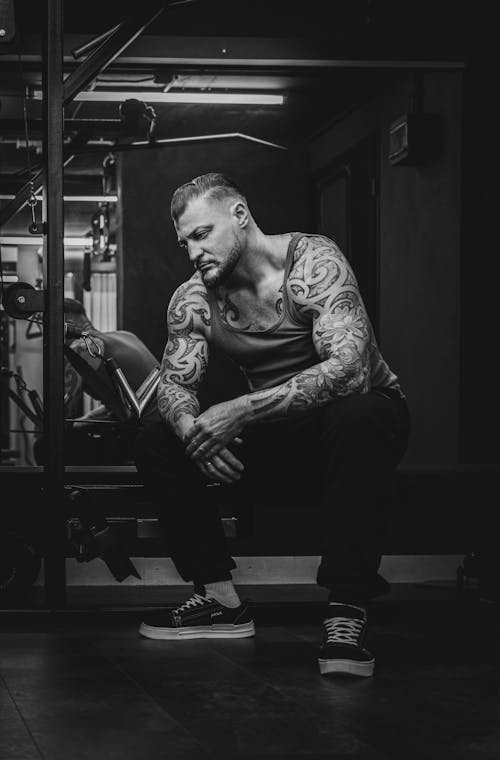Black and White Photo of a Tattooed Man in Tank Top Sitting in a Gym