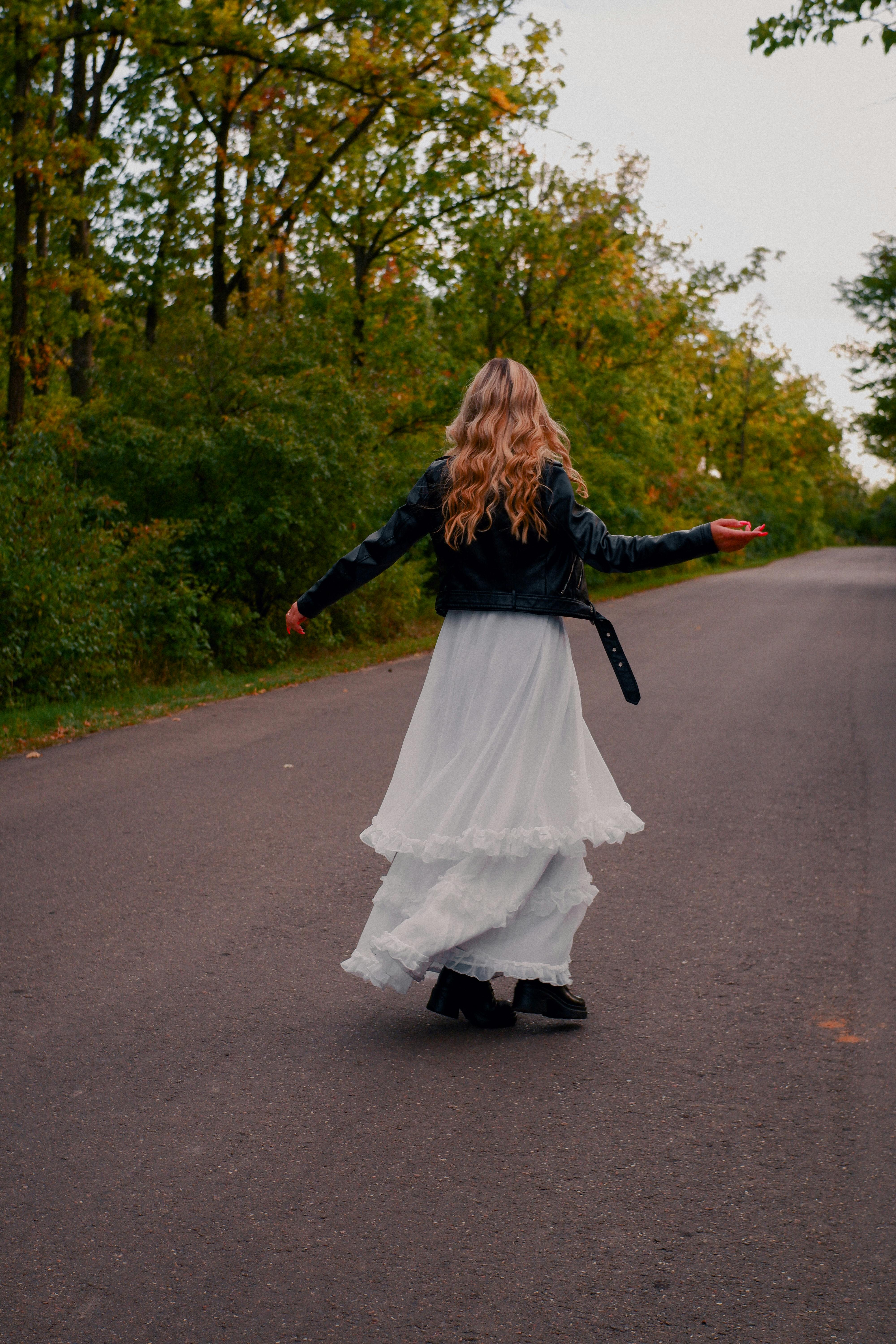 A fashionable woman twirls in a white dress with a leather jacket on a tranquil forest road.