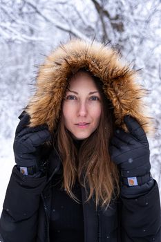 Adult woman in a fur-lined hooded coat, surrounded by a snowy winter landscape, looking directly ahead.