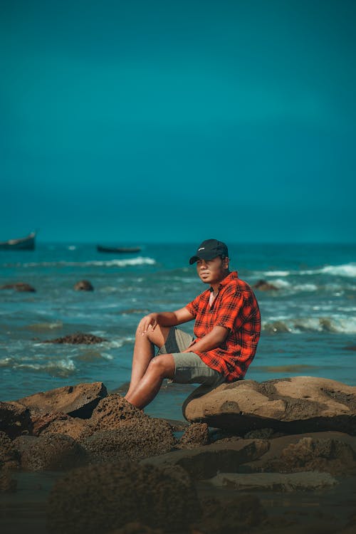 Man in a Checkered Shirt Sitting on a Rock on a Shore 