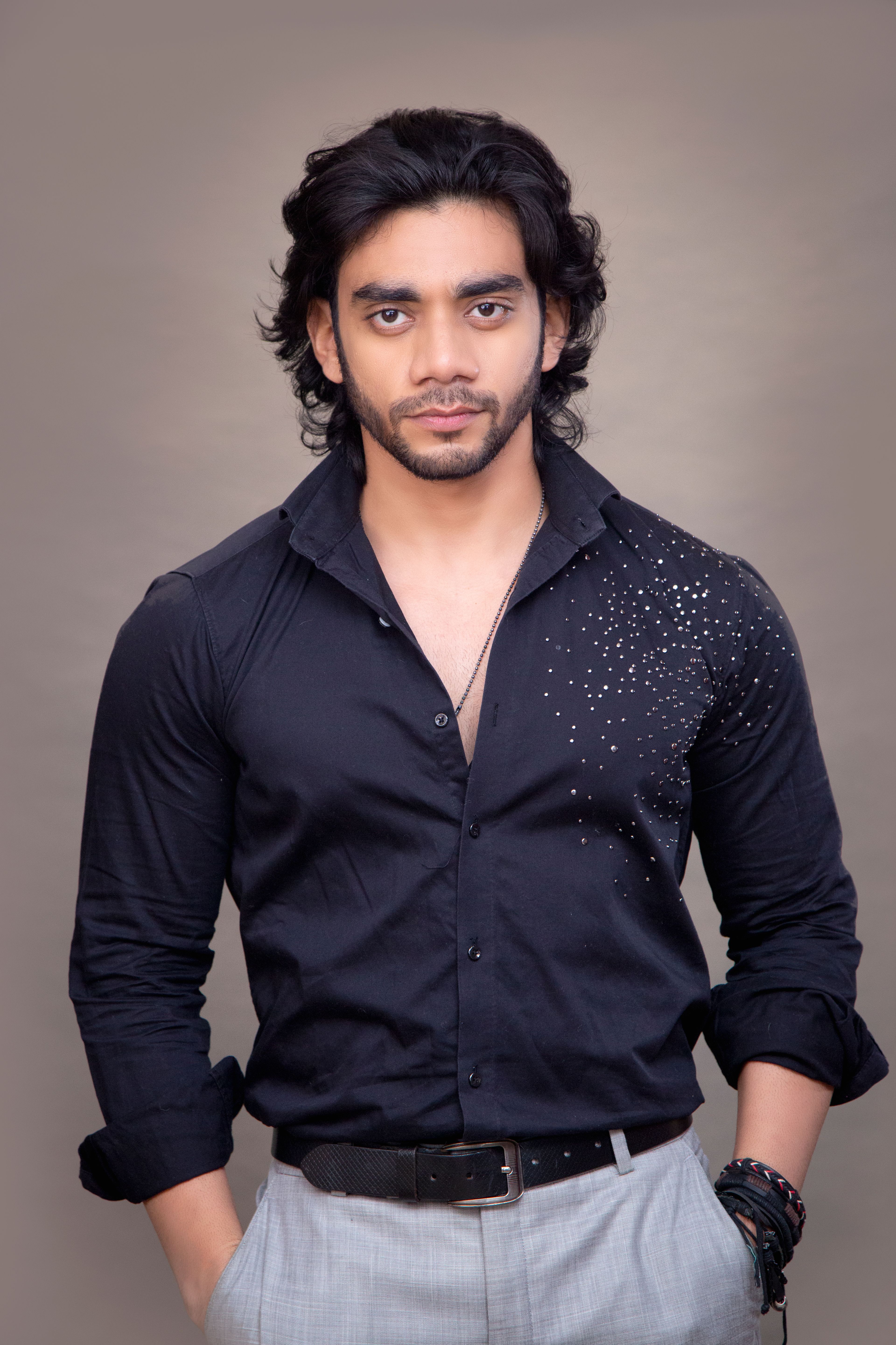 Fashionable young man posing confidently in a studio with a stylish outfit, dark hair, and accessories.