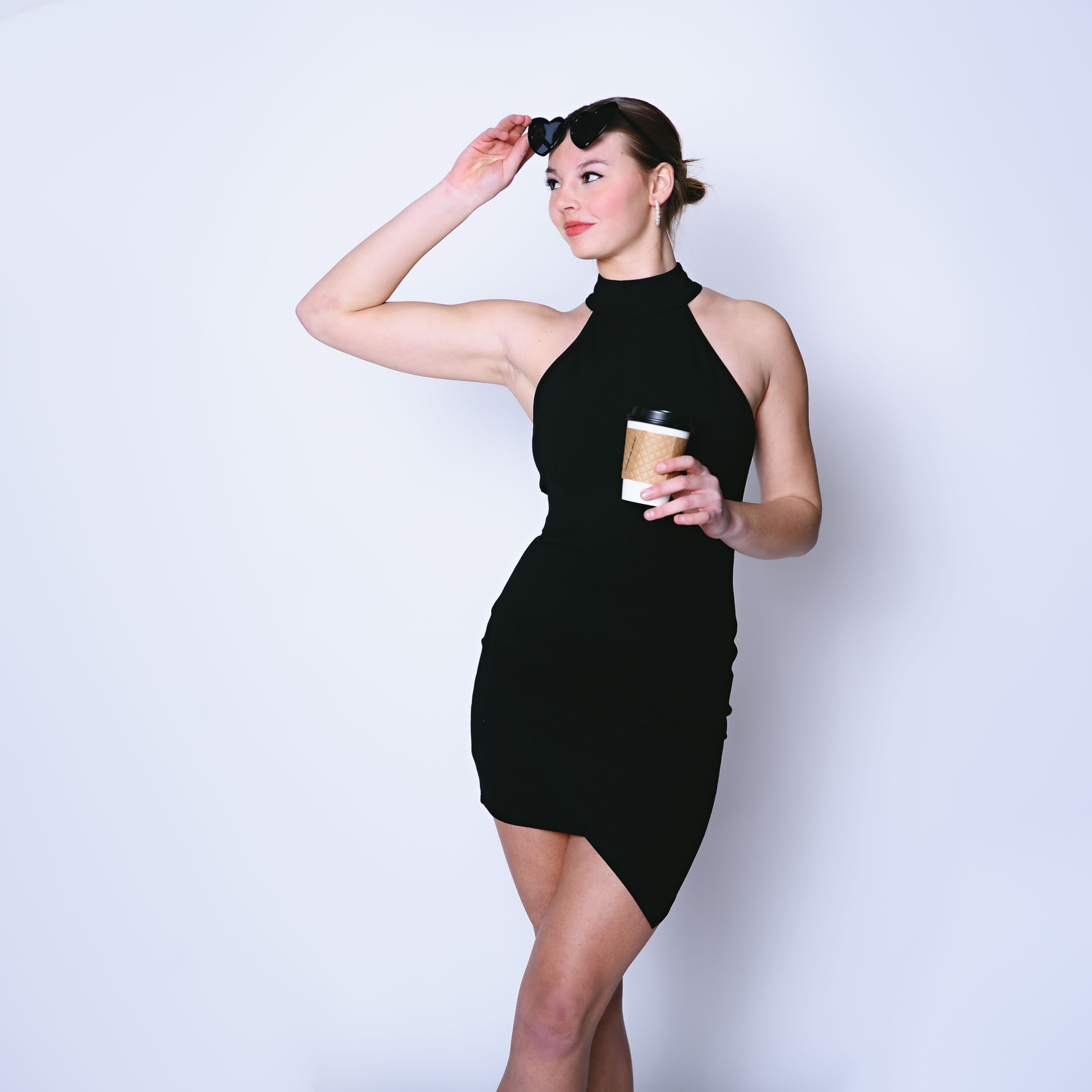 Stylish woman in a little black dress with sunglasses and coffee cup, posing confidently in a studio setting.