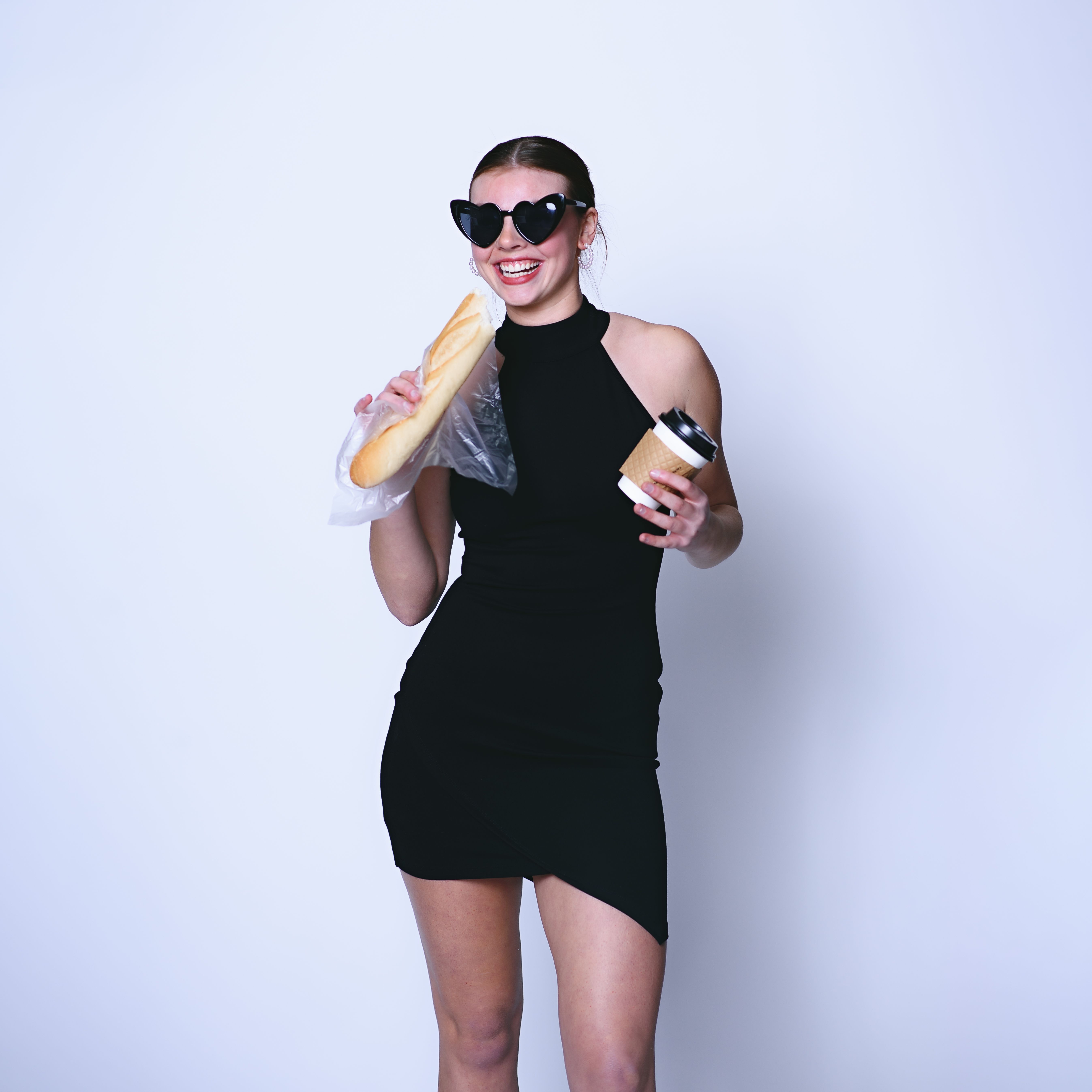 Fashionable woman in black dress with baguette and coffee, smiling on white background.