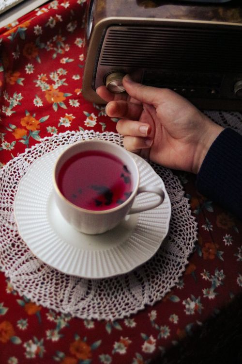 Free A hand tunes a vintage radio beside a floral tablecloth with a cup of berry tea, creating a cozy morning vibe. Stock Photo