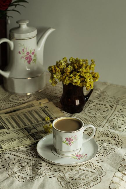 Charming floral tea set with lace tablecloth, vintage vase, and newspaper.