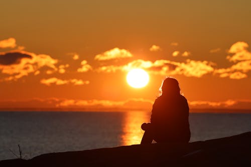 Silhouette of a Person Sitting at a Seashore and Enjoying a Sunset