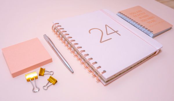 Peach-colored workspace featuring a planner, notebook, sticky notes, pen, and clips for organization.