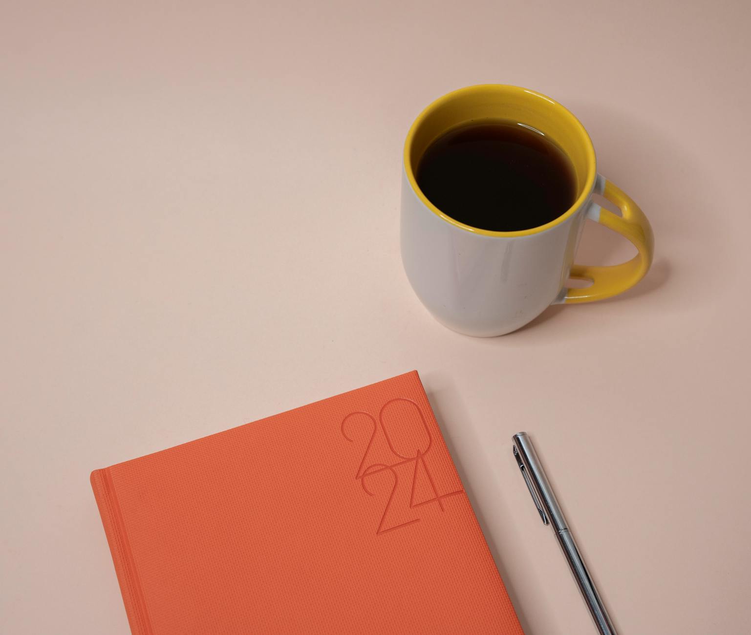 Stylish desk layout featuring a 2024 planner, cup of coffee, and pen on a light background.