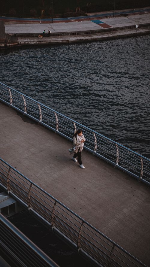 Free A solitary pedestrian walking on a riverside walkway, captured in an urban environment with water and bridge structures. Stock Photo