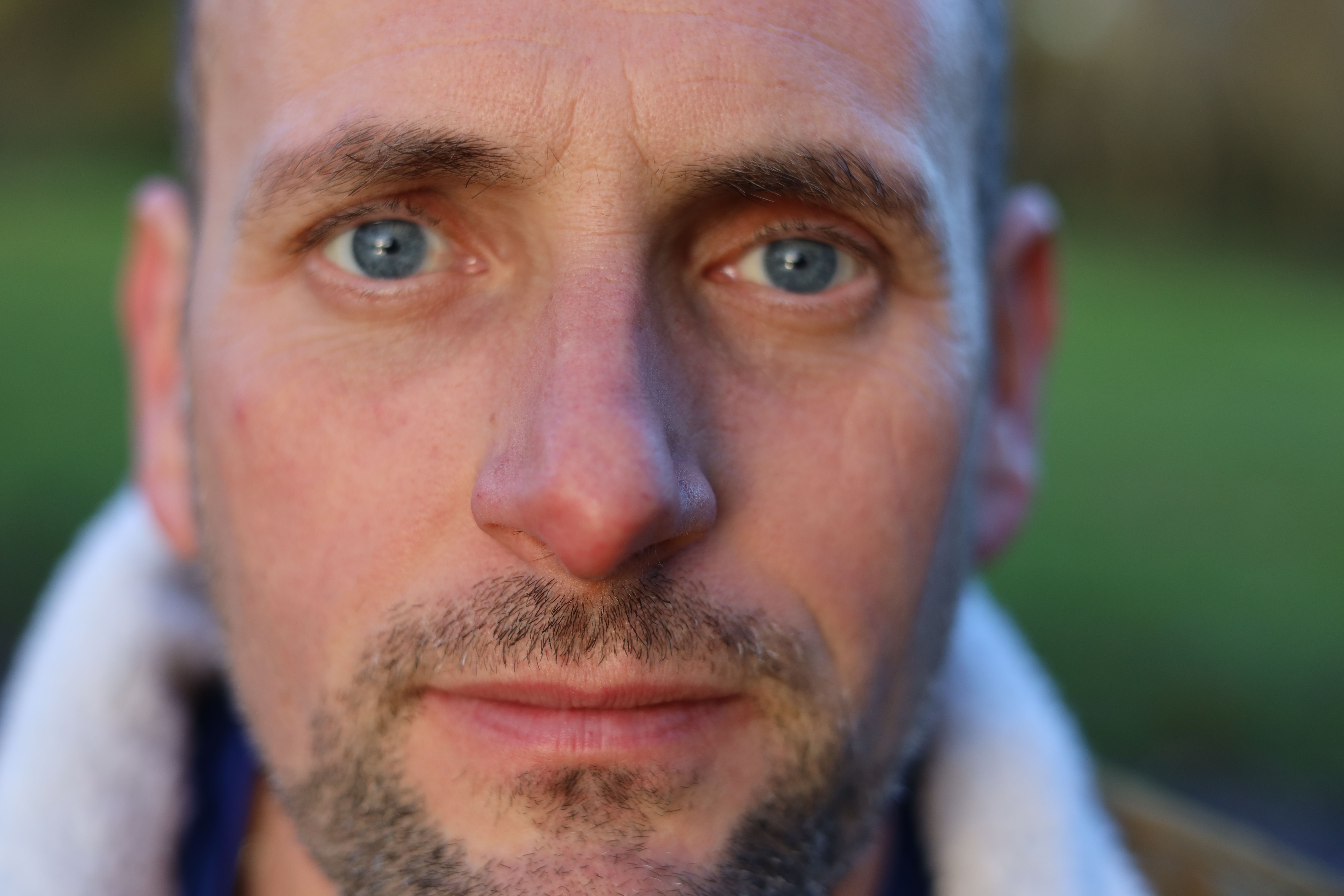 A close-up portrait of a serious man with blue eyes in an outdoor setting.