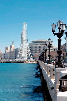 A scenic view of a Ferris wheel along the urban coastline with blue sea and street lamps.