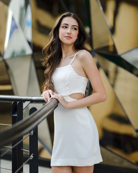 Stylish brunette woman wearing a white dress poses elegantly outdoors in a modern urban setting.