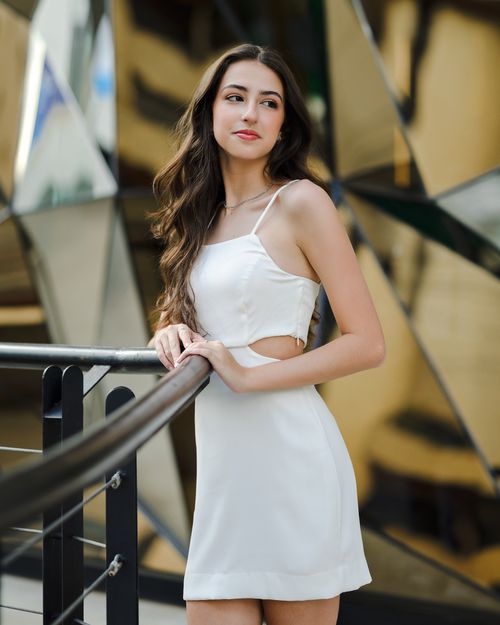 Free Stylish brunette woman wearing a white dress poses elegantly outdoors in a modern urban setting. Stock Photo