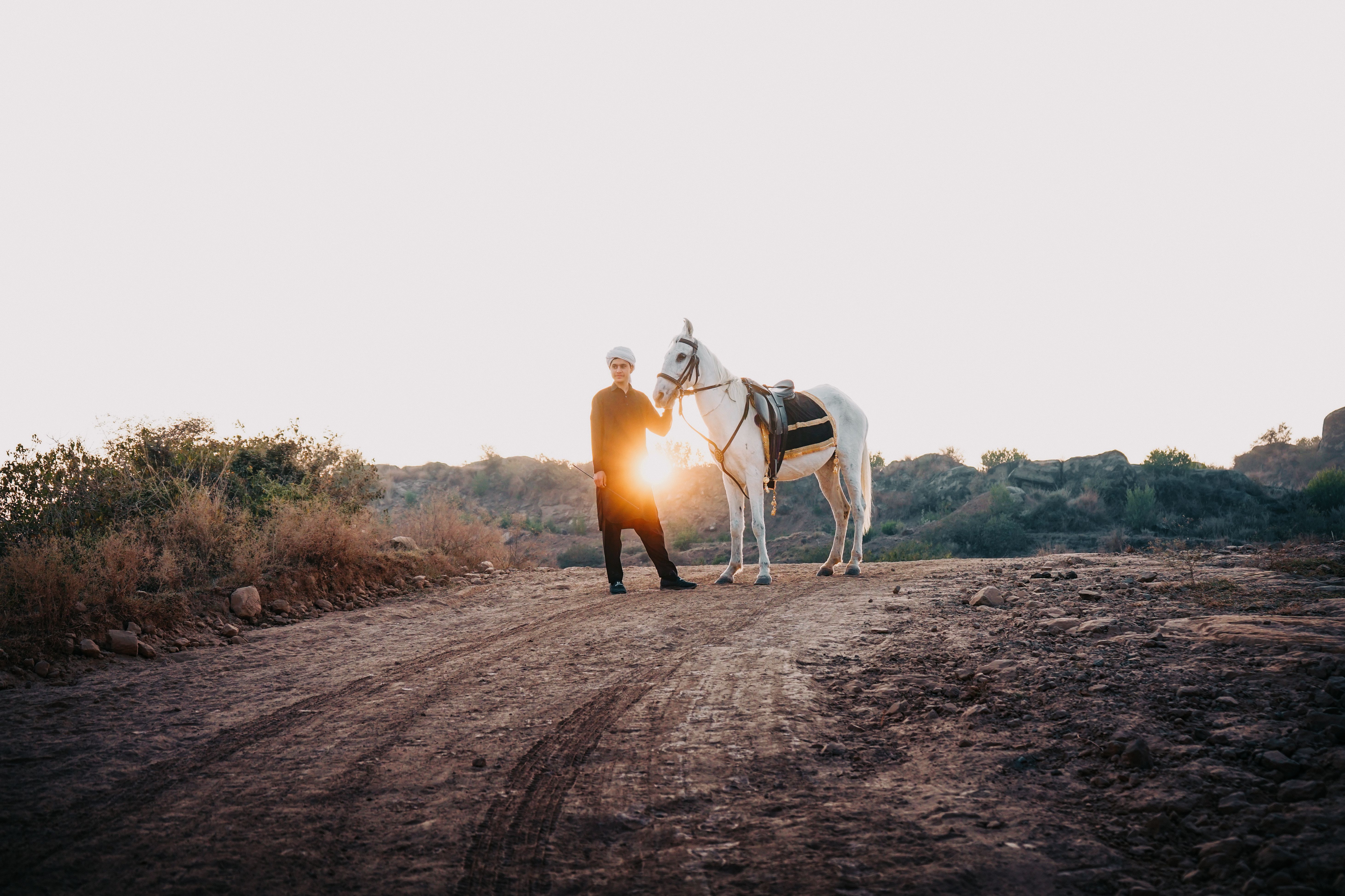 A man in traditional clothing stands with a white horse on a rural dirt road during sunset, capturing a serene countryside scene.