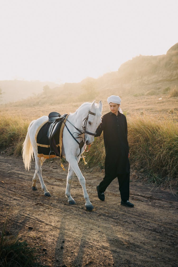 Man Leading A Saddled White Horse Along A Dirt Road