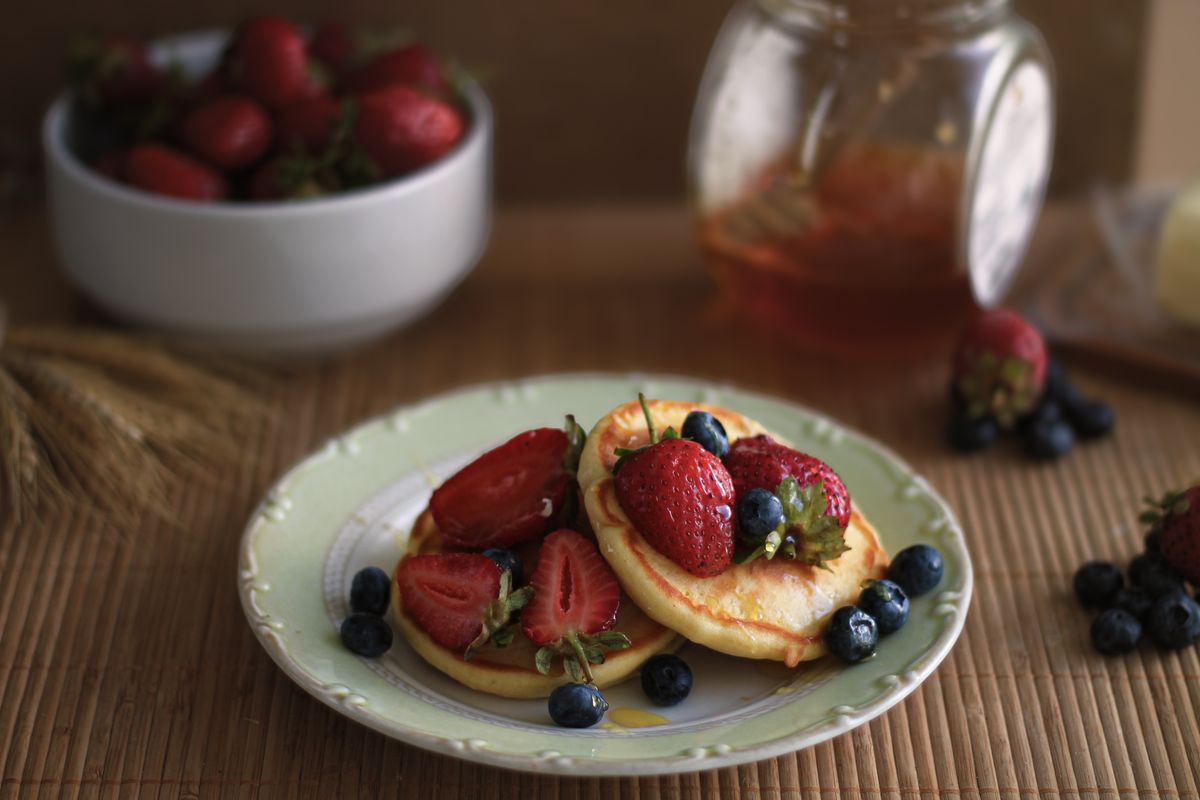 Small fluffy pancakes topped with halved strawberries and blueberries beside a honey jar
