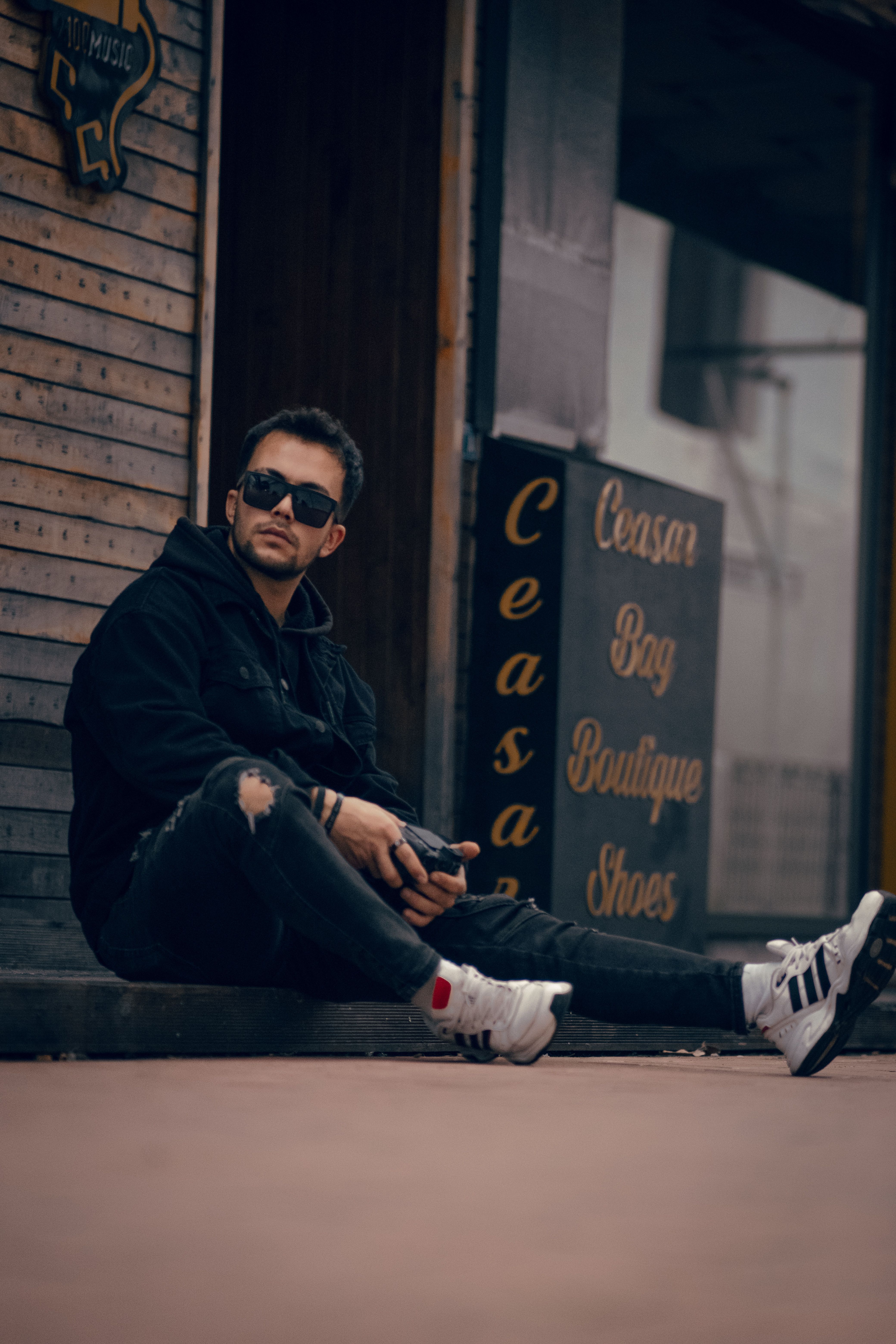 Stylish man in sunglasses sitting on city sidewalk with urban fashion vibe.