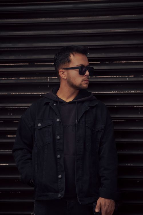 Free A young man in a black hoodie and denim jacket wearing sunglasses, standing against a roller door. Stock Photo
