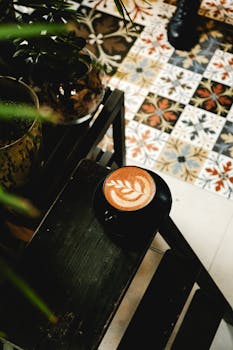 A beautifully crafted latte with intricate leaf art, placed in a cozy cafe with vibrant tiled flooring.