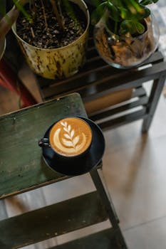 Fresh cappuccino with elegant latte art on a wooden table in a cozy café, surrounded by greenery.