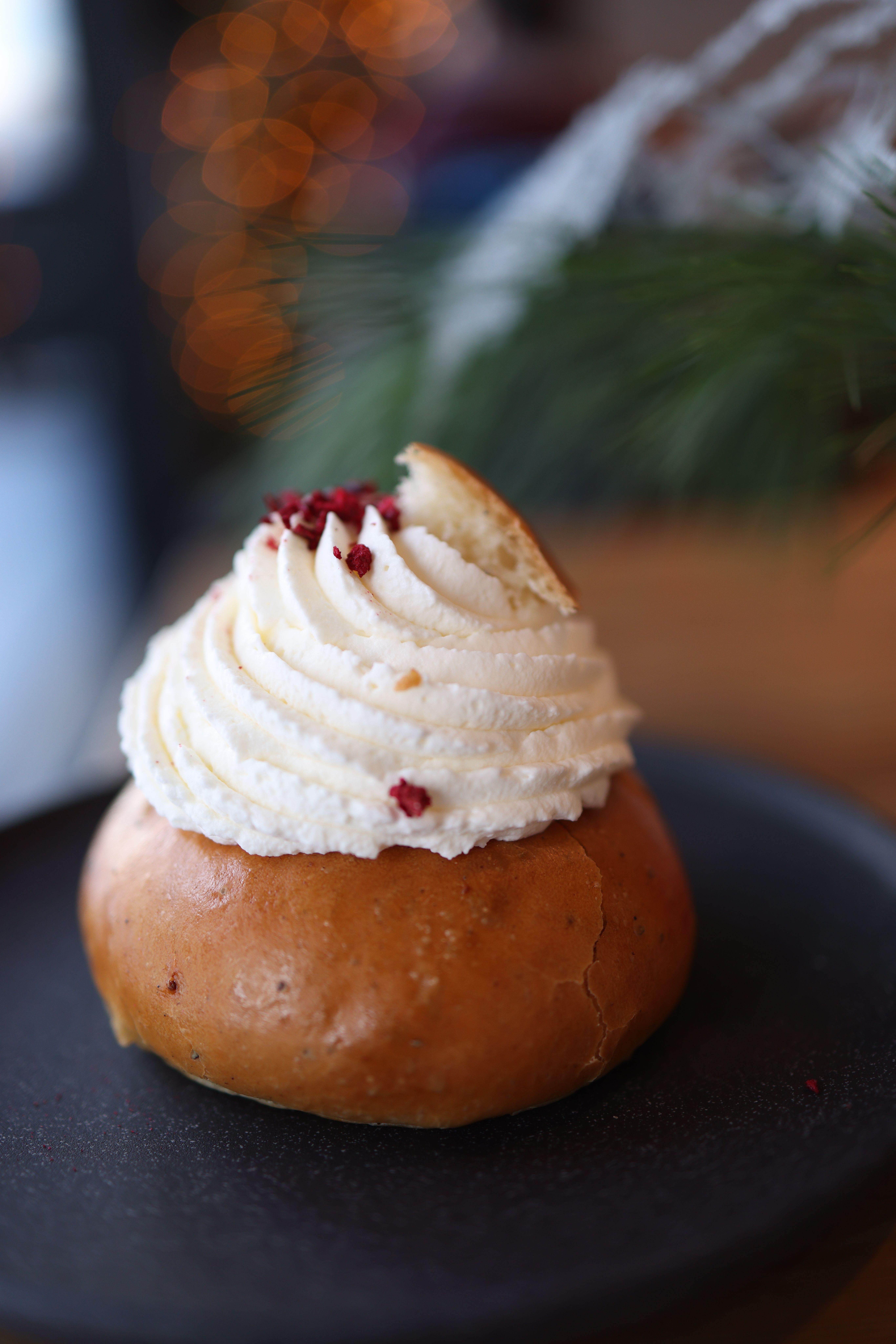 Close-up of a fresh pastry bun topped with whipped cream and sprinkles, set on a dark plate.