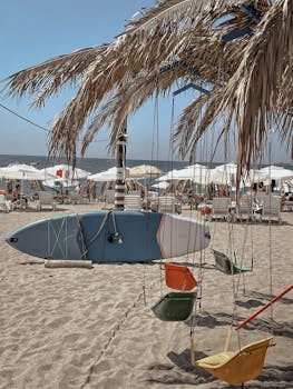 Sunny beach with swings, surfboard, and chairs under palm trees, perfect for a tropical vacation.