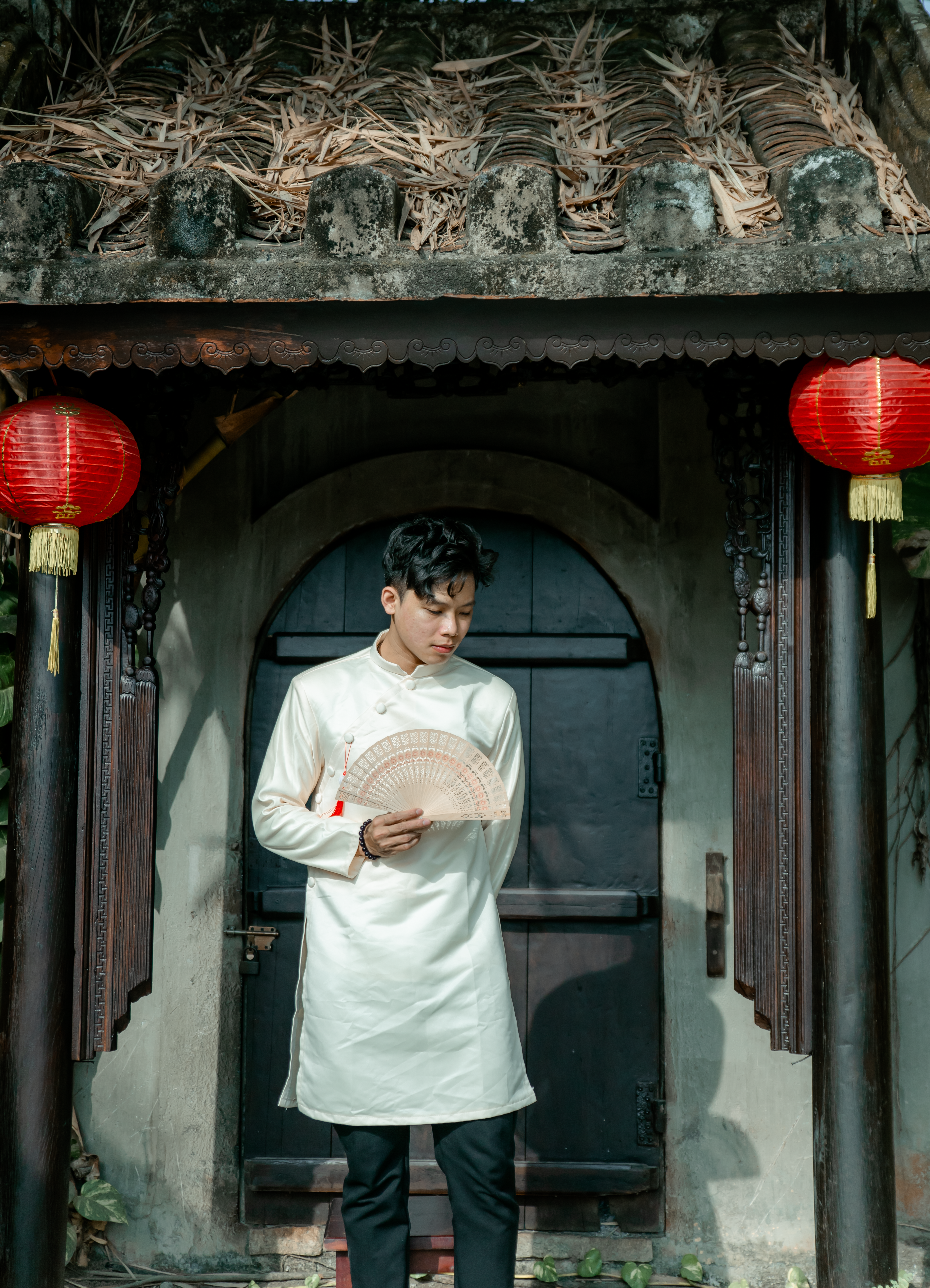 A young man in traditional clothing stands by a door with red lanterns, holding a fan.