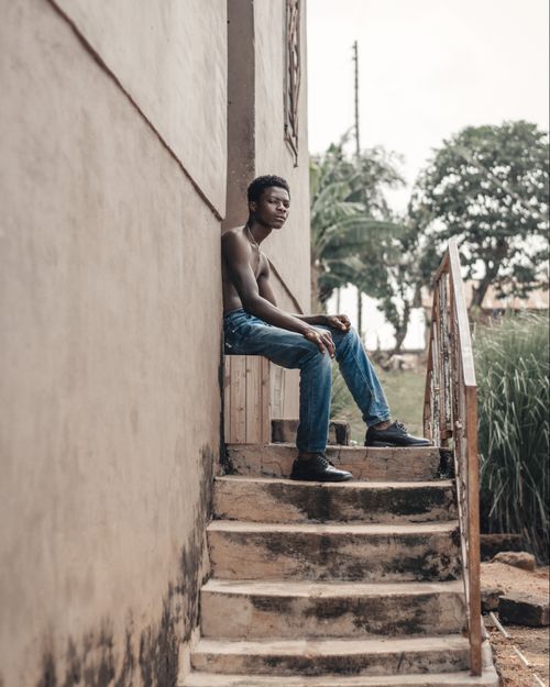 Free A topless young man sits on concrete steps in a rustic village setting, conveying a calm and reflective mood. Stock Photo