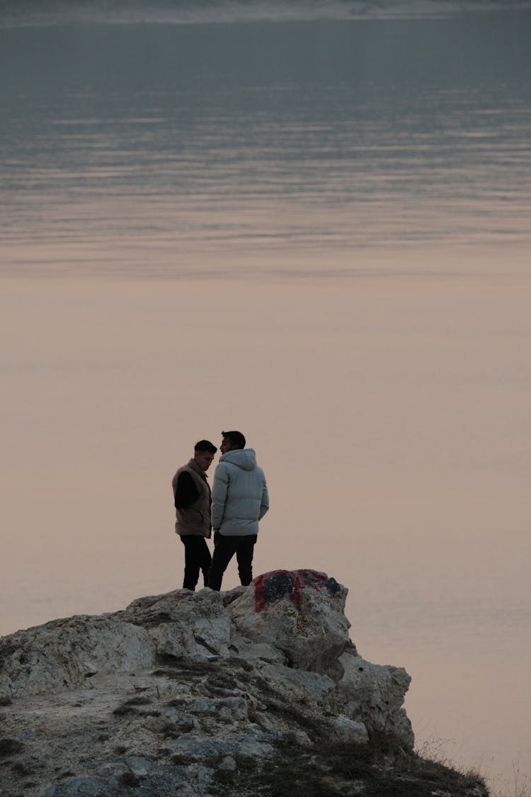 Two Men Standing On A Rocky Hill Overlooking A Lake At Dusk