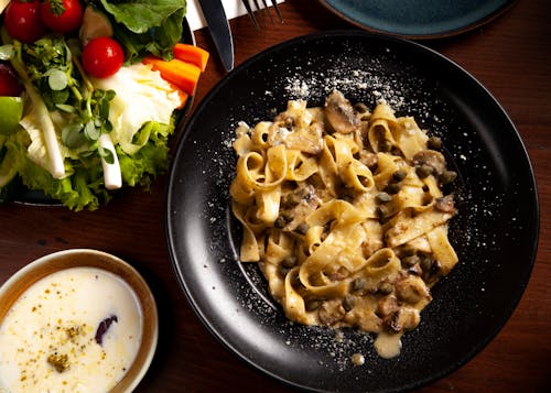 Pasta with Mushrooms on a Black Plate, a Bowl of Salad and a Dipping Sauce