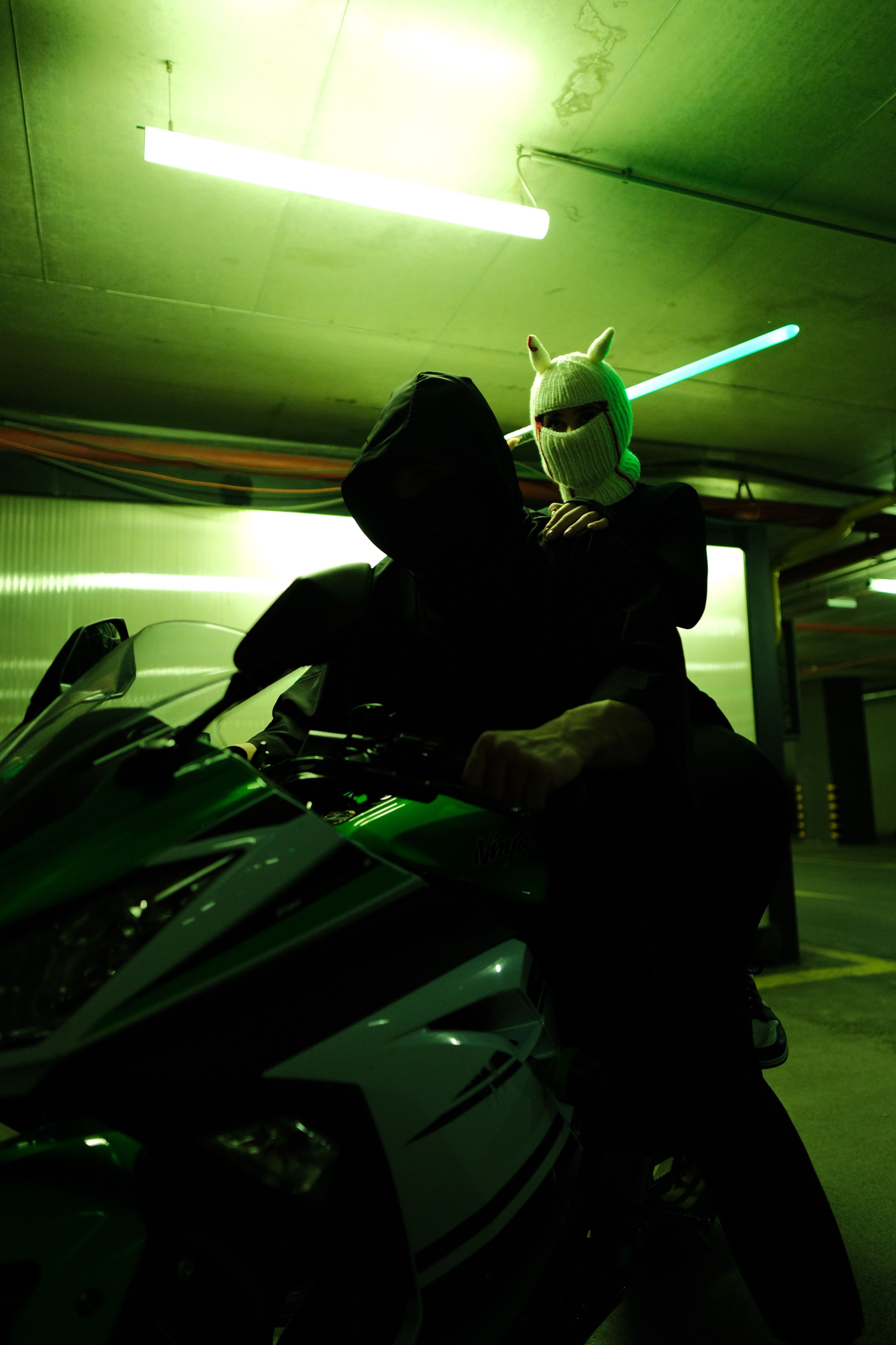 Two figures on a modern motorcycle in a neon-green-lit urban garage.