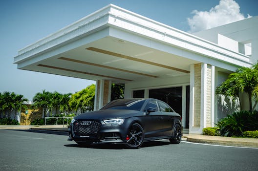 A sleek black Audi RS3 sports car parked in front of a modern building with clear blue skies and lush greenery.