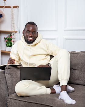 A smiling man in a yellow tracksuit sits on a sofa with a laptop and notebook, indoors.