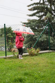 A child in a red jacket holding an umbrella stands on wet grass by a fence, with trees in the background.