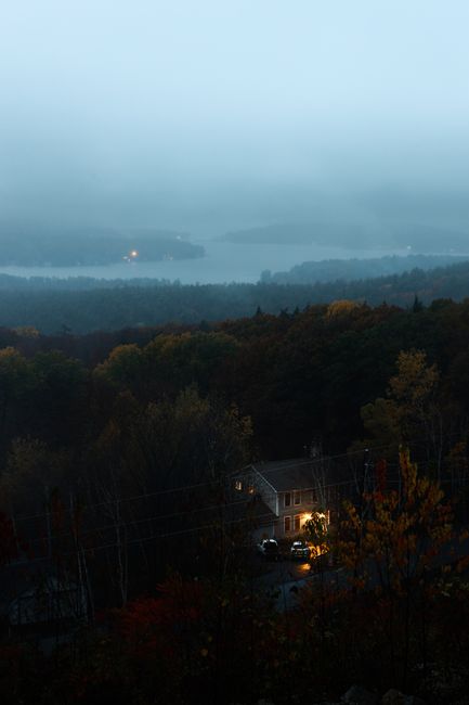 Aerial view of a forest landscape at dusk with a glowing house, surrounded by misty mountains and trees.