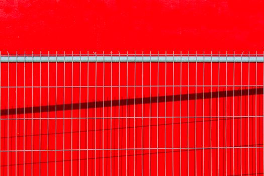 A vibrant red wall with a metal fence casting shadows, creating a striking abstract pattern.