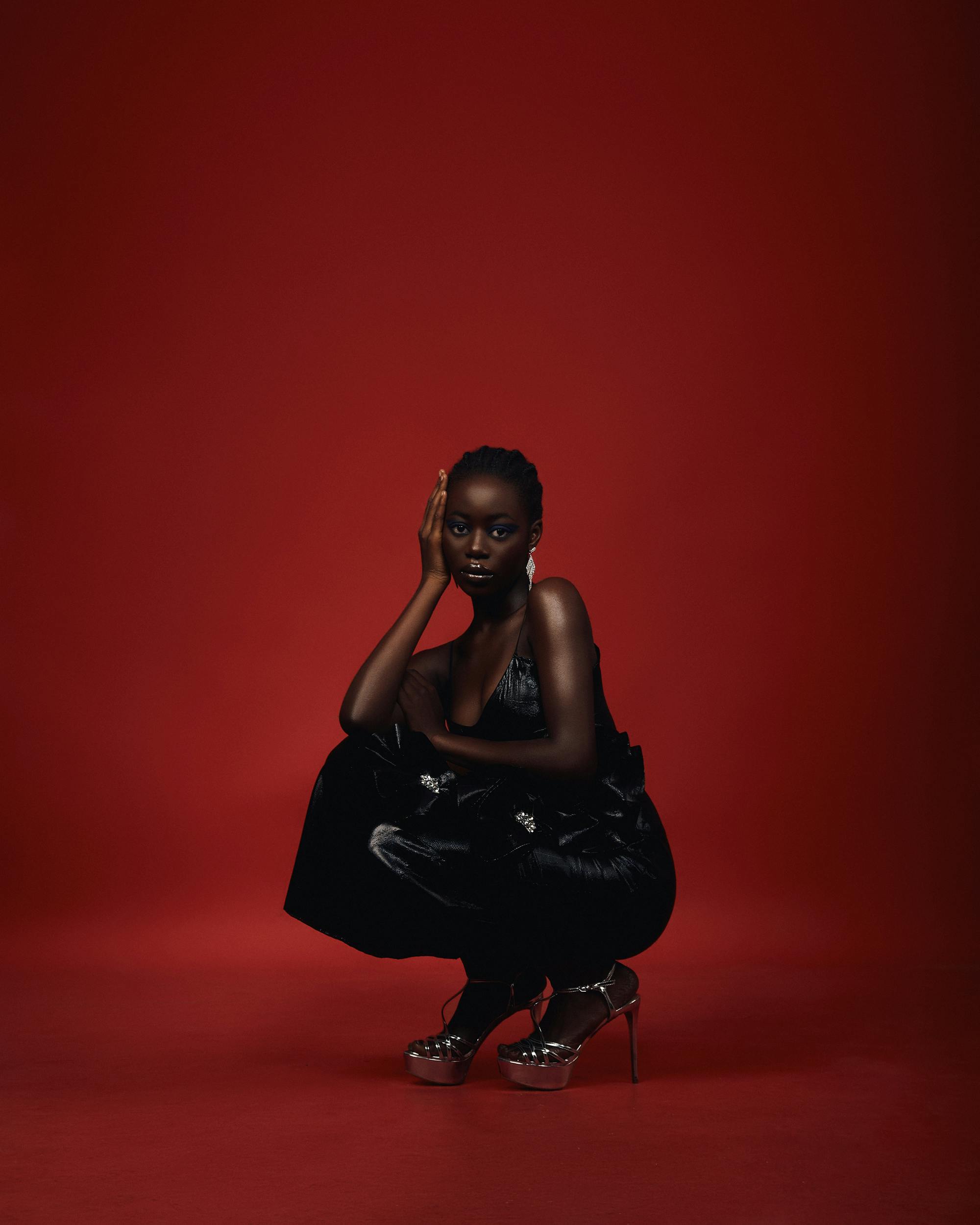 Portrait of a model in a black dress squatting on a striking red background, embodying elegance.