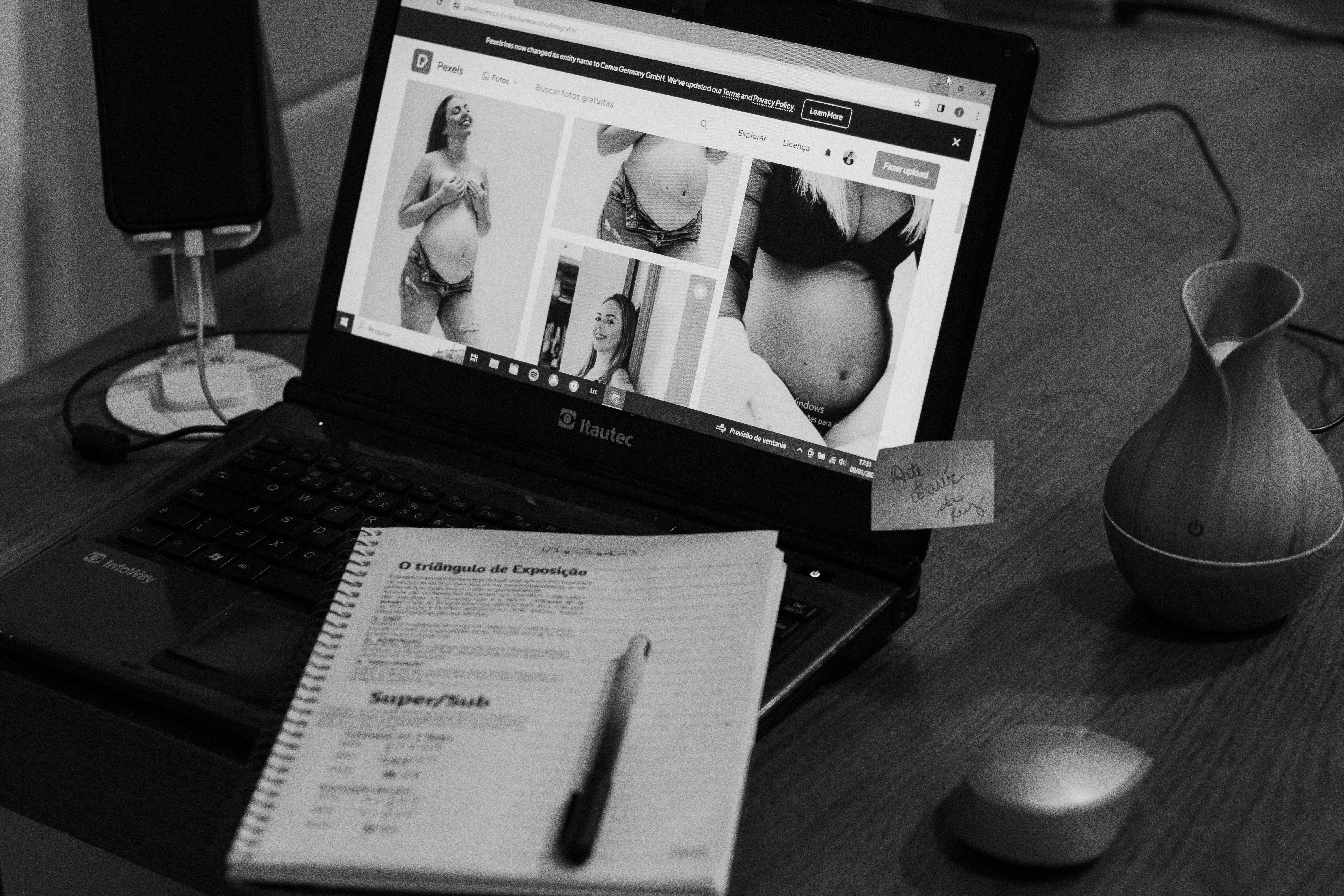 Black and white photo of a laptop showing pregnancy images with a notepad and pen beside it.