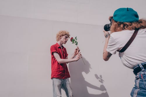 Woman Taking Photo of Man Standing and Holding Red Flower Near Wall