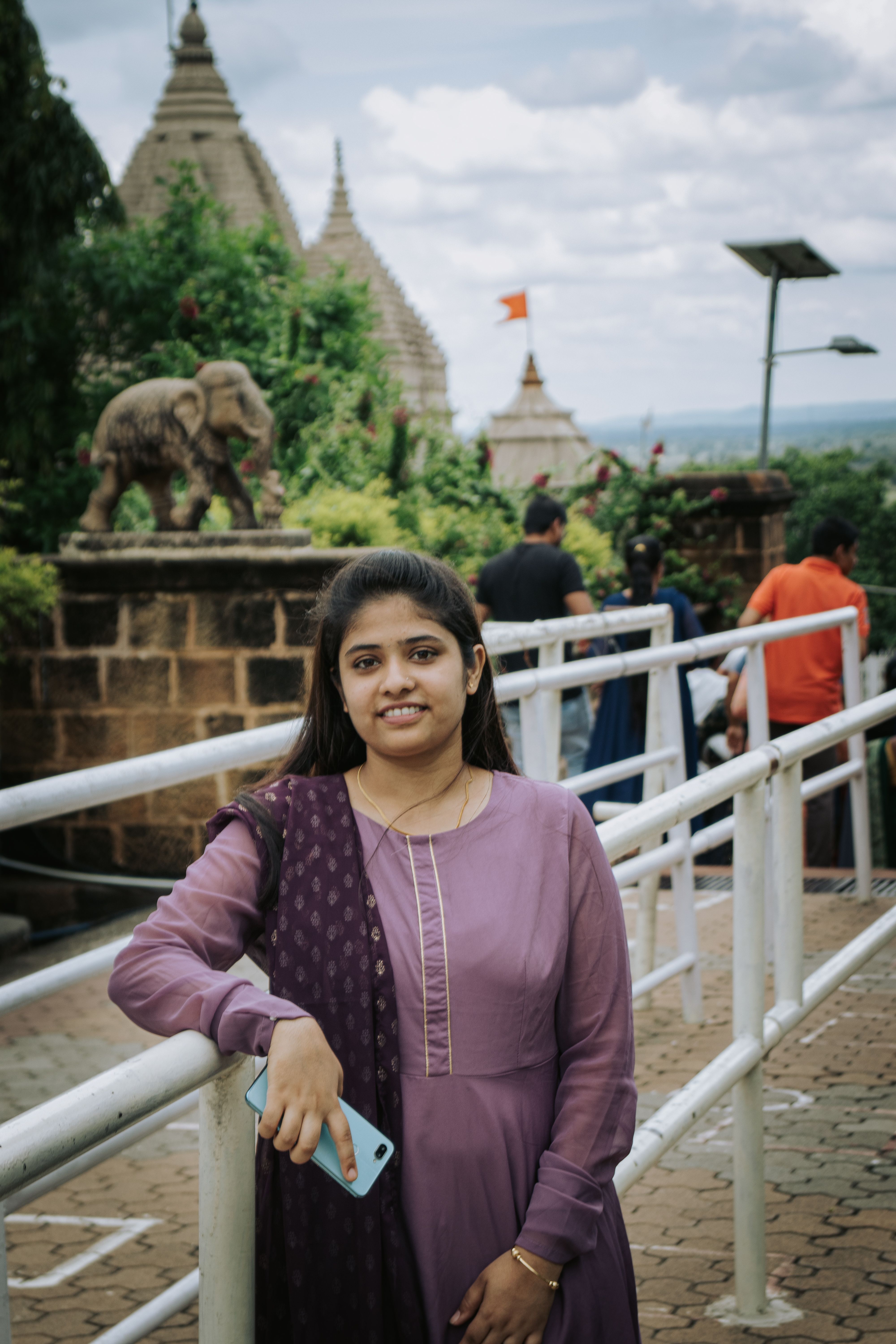 A smiling young South Asian woman in a purple dress stands near a temple.