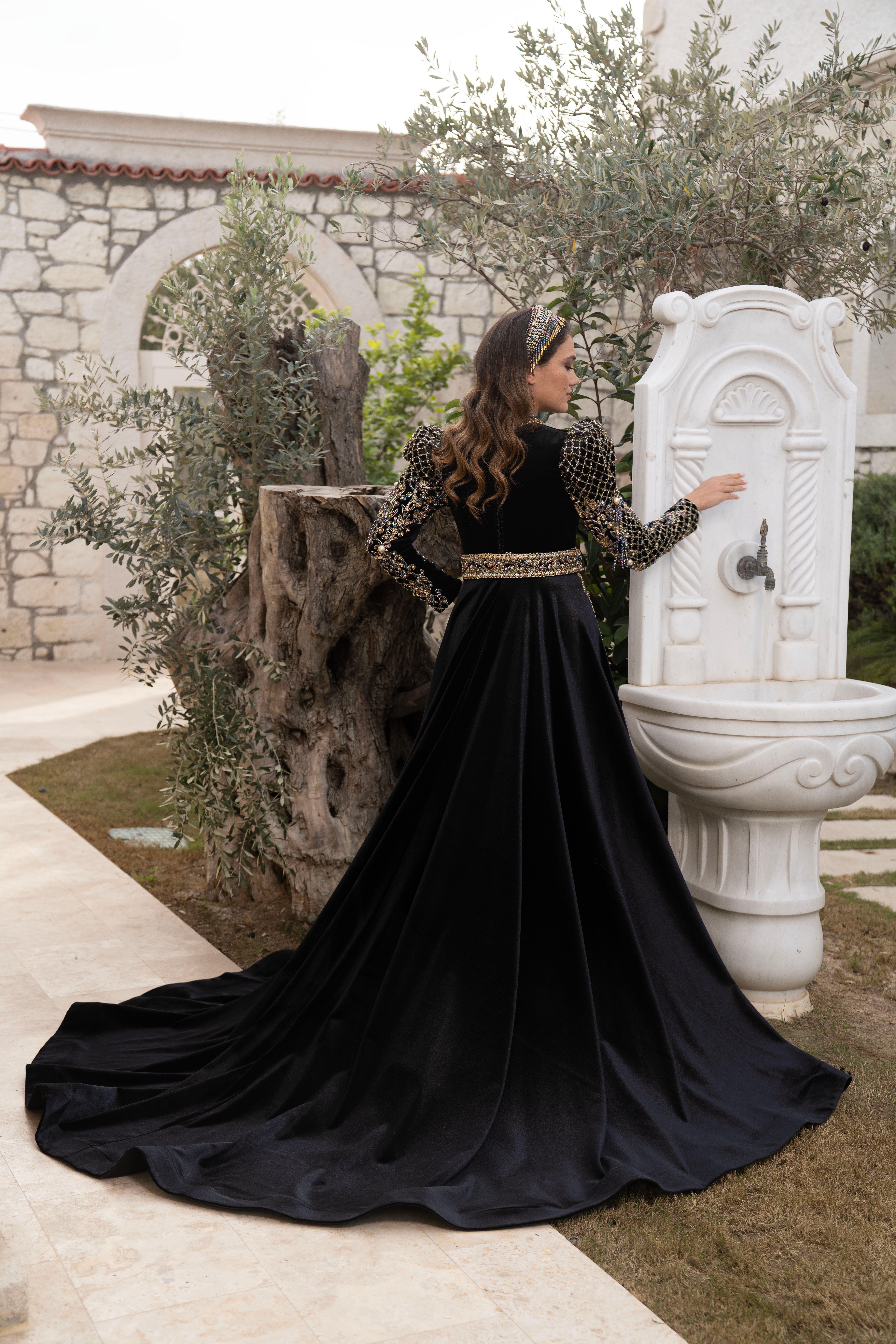 A woman wearing a luxurious black velvet dress poses by a vintage fountain outdoors in Izmir.