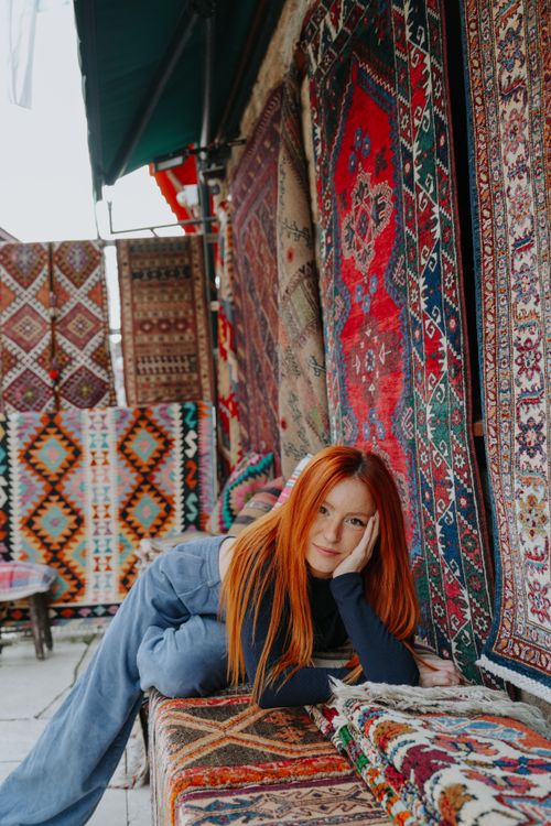 Free A red-haired woman in stylish attire lies among colorful patterned rugs, creating a vivid and artistic scene. Stock Photo